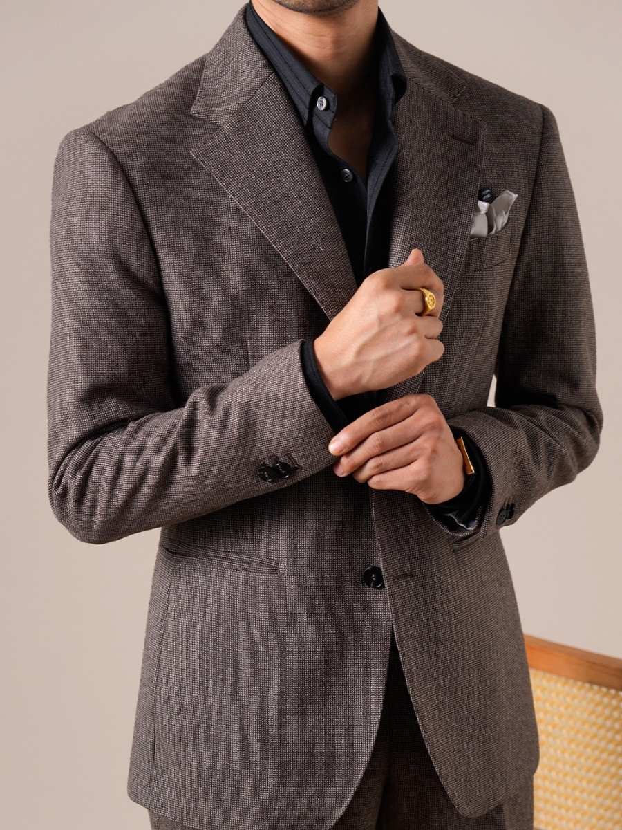 Man wearing tailored brown checkered suit with black shirt, adjusting sleeve, gold ring and watch visible