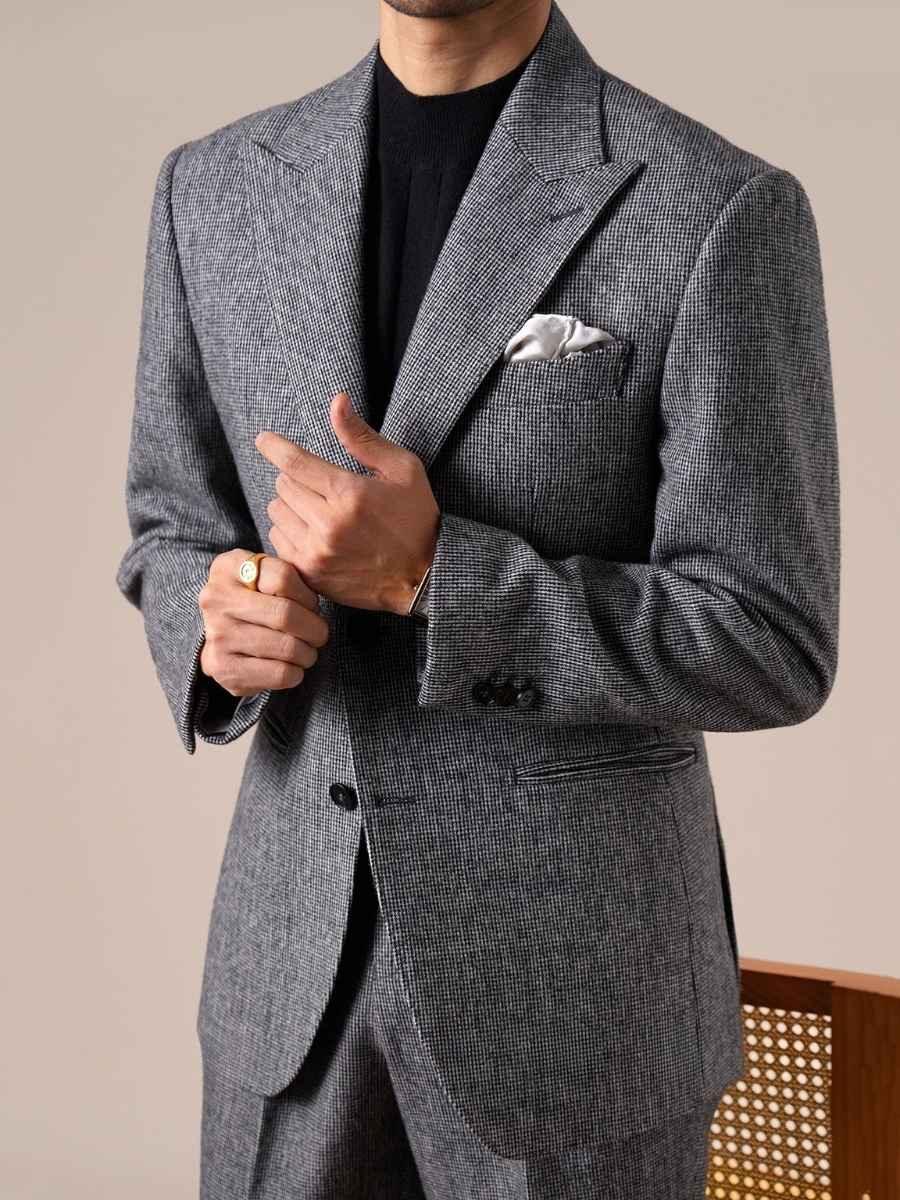 Man wearing a grey checkered suit with a white pocket square and black shirt, adjusting his jacket