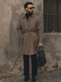 Man in brown checkered belted coat, black sunglasses, black pants, holding black leather bag against worn wall