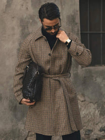 Man in houndstooth coat, black turtleneck, sunglasses, and gold wristwatch holding black woven leather bag